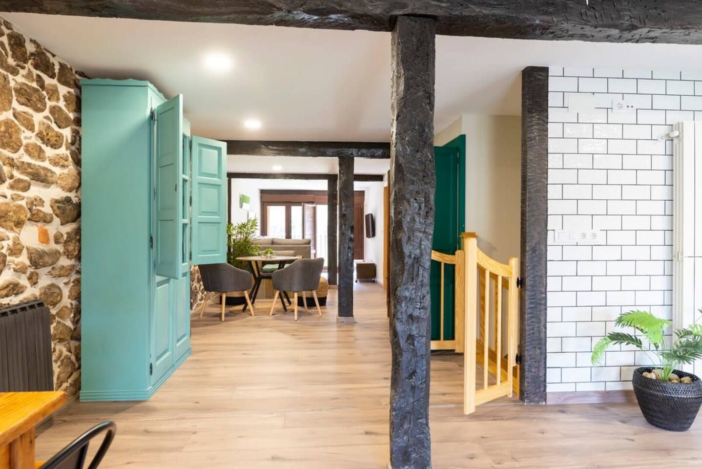 A room with a stone wall, a teal cabinet, a wooden floor, a dining area with a table and chairs, and a staircase with a wooden railing.