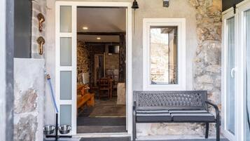 Fachada de piedra con un banco, una puerta que da a una habitación con chimenea y una ventana.