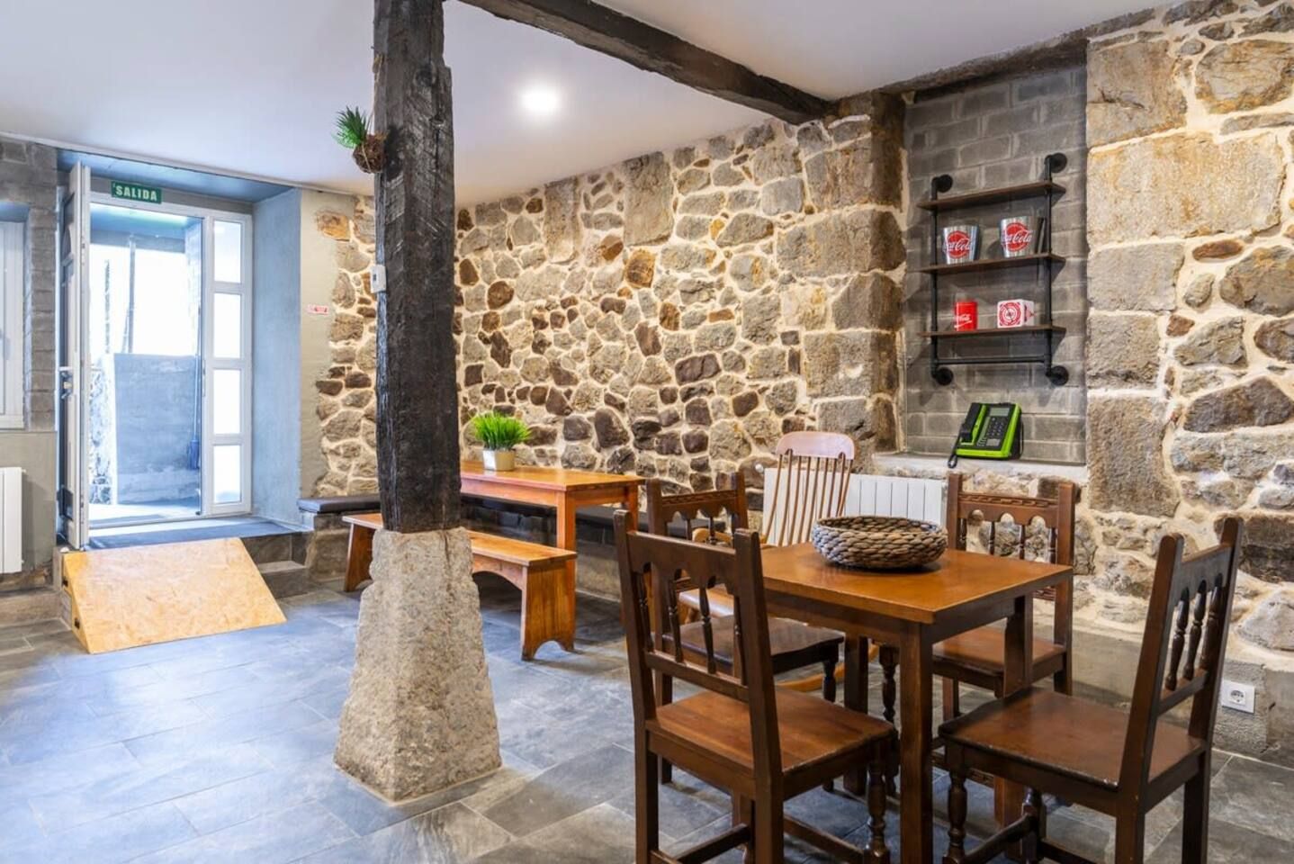 Una habitación con paredes de piedra, una mesa de madera y sillas, un banco y una puerta con la etiqueta 'SALIDA'.