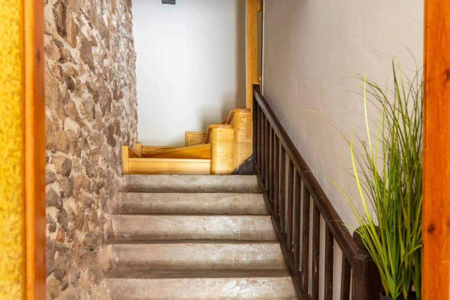 Una escalera con un muro de piedra a un lado y barandilla de madera al otro, que conduce a una habitación con puerta blanca.