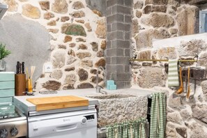 A kitchen with a stone wall, a wooden cutting board, a white dishwasher, and a hanging towel rack. - El Cason Riva Ruesga (Riva)