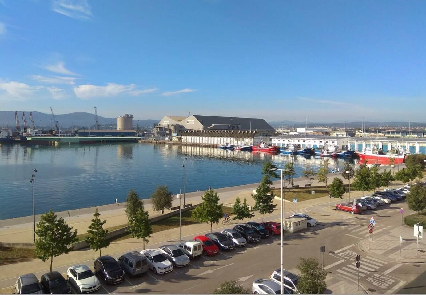 Un puerto deportivo con barcos amarrados, un estacionamiento con coches y edificios industriales al fondo.
