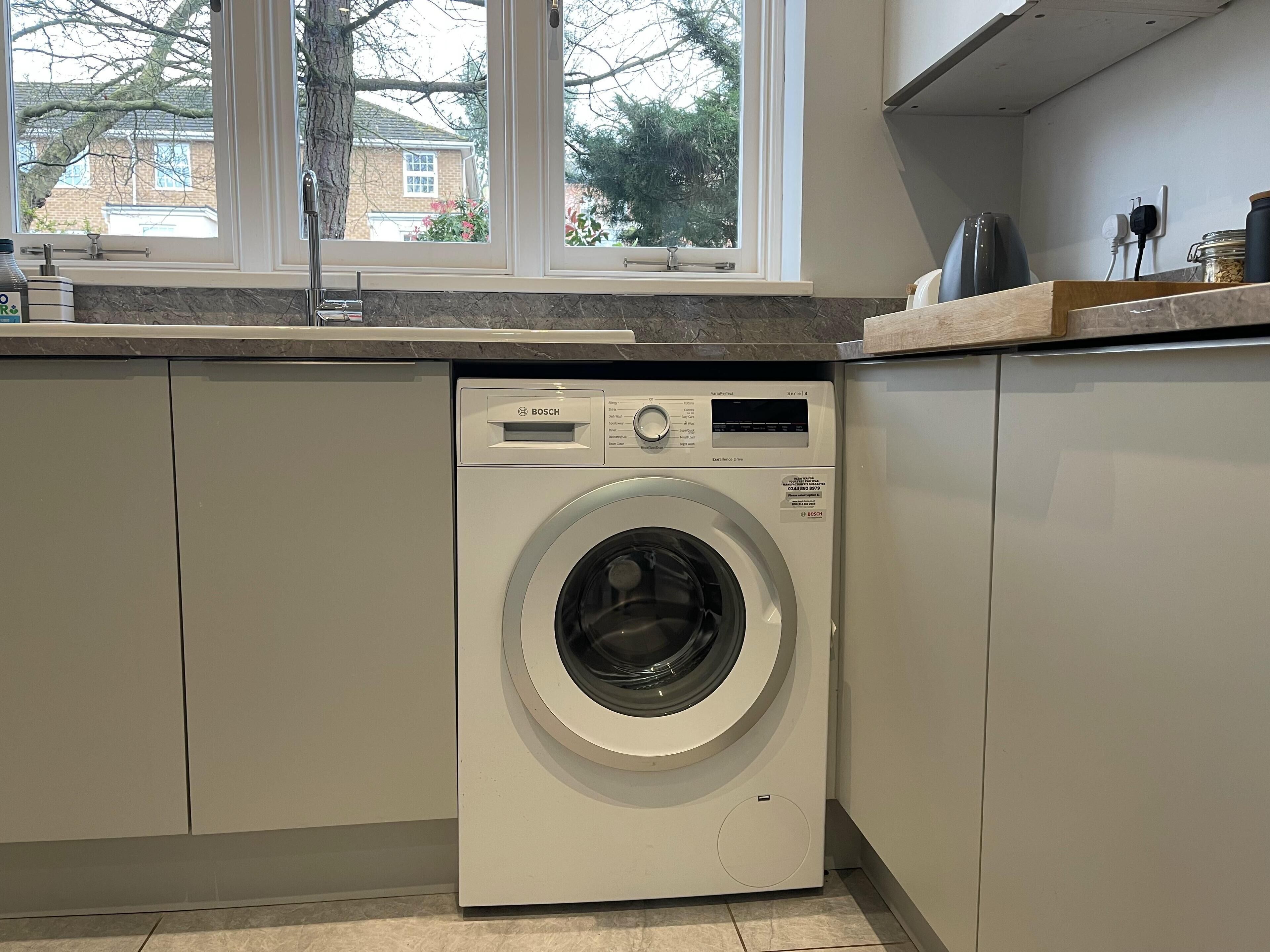 A compact, white Bosch washing machine in a modern kitchen with a window above the sink.
