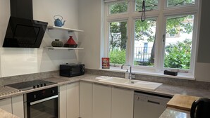 A modern kitchen with a built-in oven, microwave, and dishwasher, a window with a view of greenery, and a shelf with decorative items. - 1bedapt Privateestate W/free Parking - 10mintocity (Nottingham)