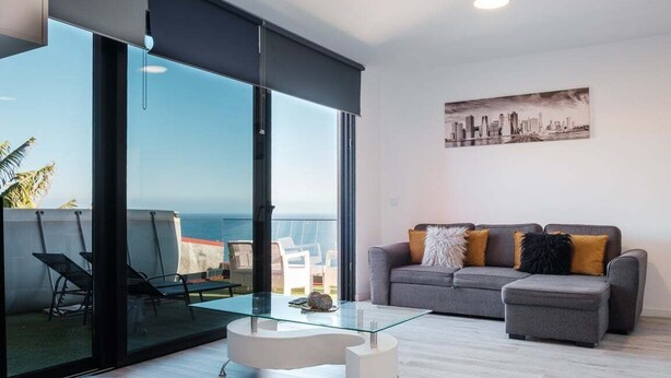 A modern living room with a glass coffee table, grey sofa, and a cityscape painting. - GuestReady - Sonho dos Avós I (Câmara de Lobos)