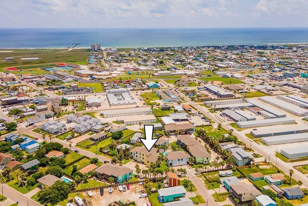 An aerial view of a coastal town with residential and commercial buildings, a sports field, and a view of the ocean.
