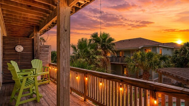 A wooden deck with chairs, a clock, and string lights against a sunset backdrop.