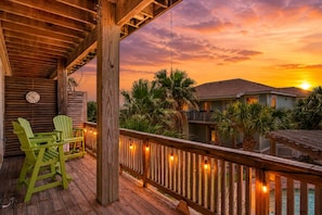 A wooden deck with chairs, a clock, and string lights against a sunset backdrop. - Casa Del Mar - Golf Cart Included! (Port Aransas)