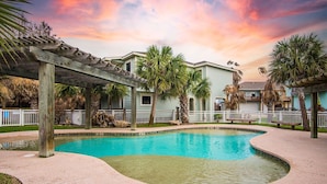 A swimming pool with a wooden pergola, palm trees, and a house in the background.
