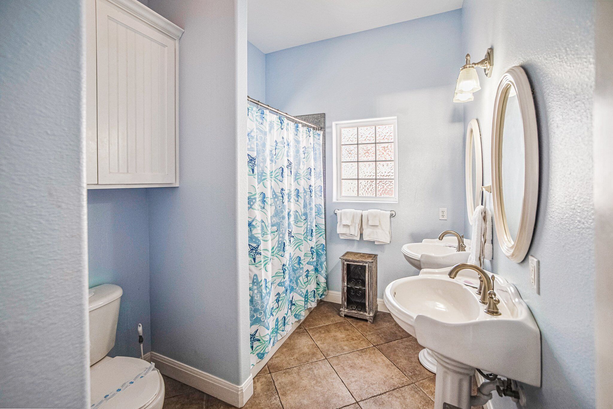 A bathroom with a toilet, a shower with a butterfly shower curtain, two sinks, a window, and a mirror.
