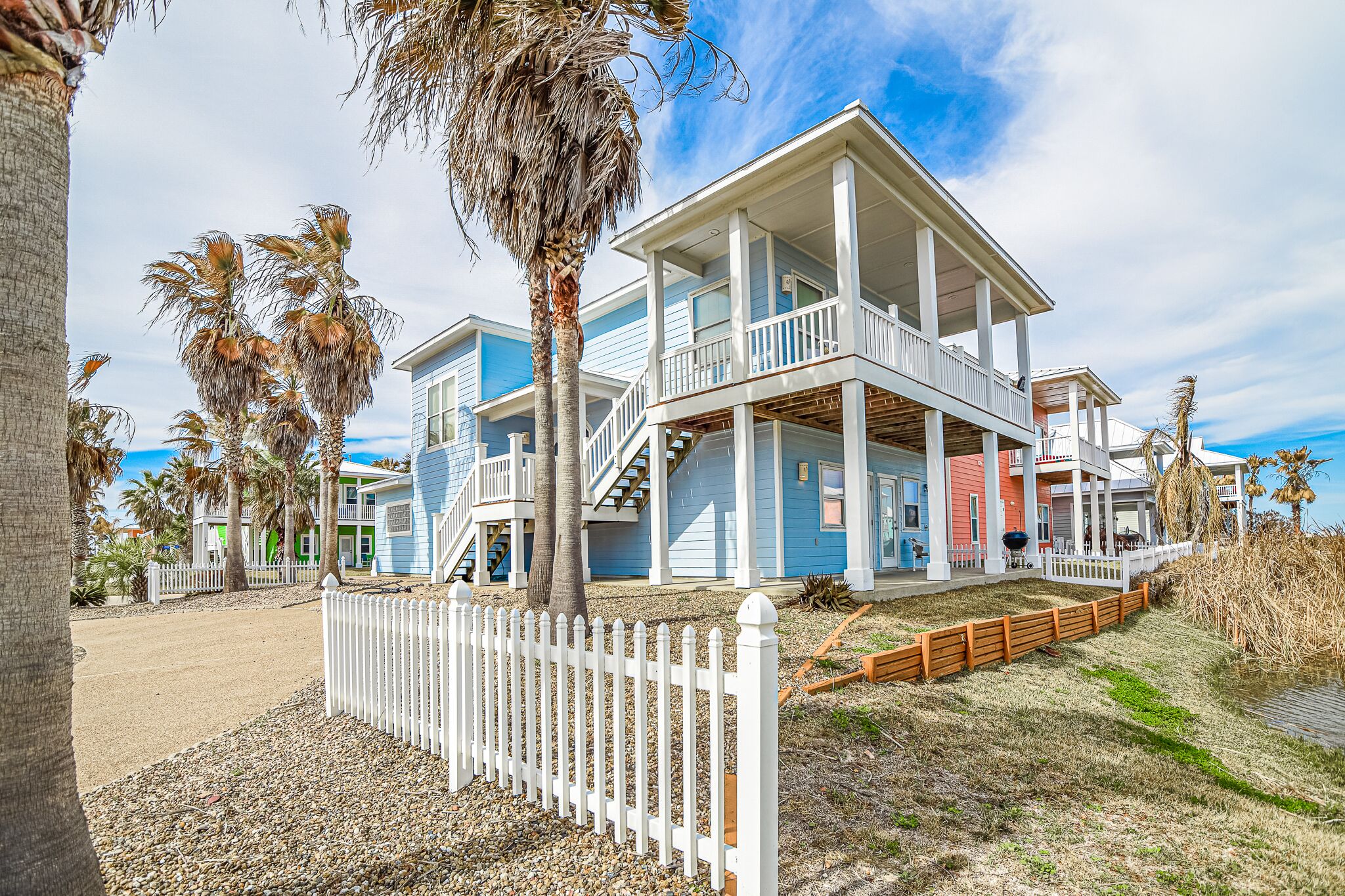 A beachfront property with multiple levels, a white picket fence, and palm trees.