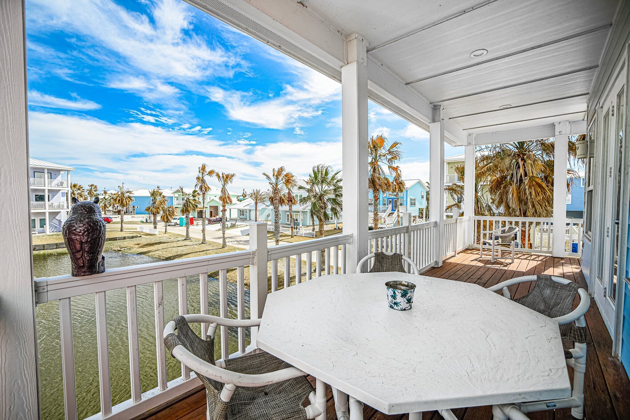 A covered outdoor area with a white table and chairs, a decorative bird sculpture, and a view of palm trees and buildings across a body of water.