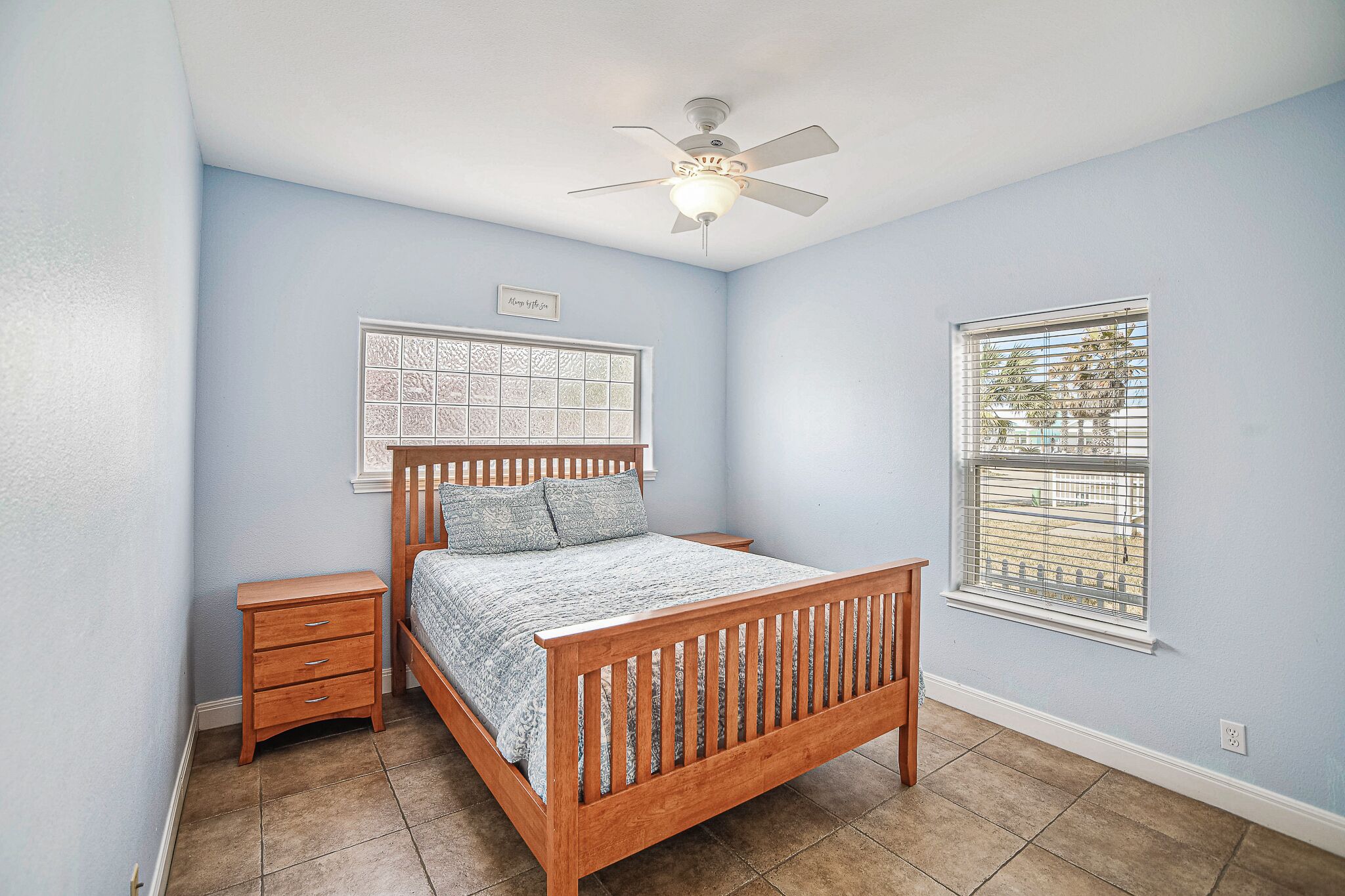 A bedroom with a wooden bed, a nightstand, a ceiling fan, a window with blinds, and a decorative wall panel.