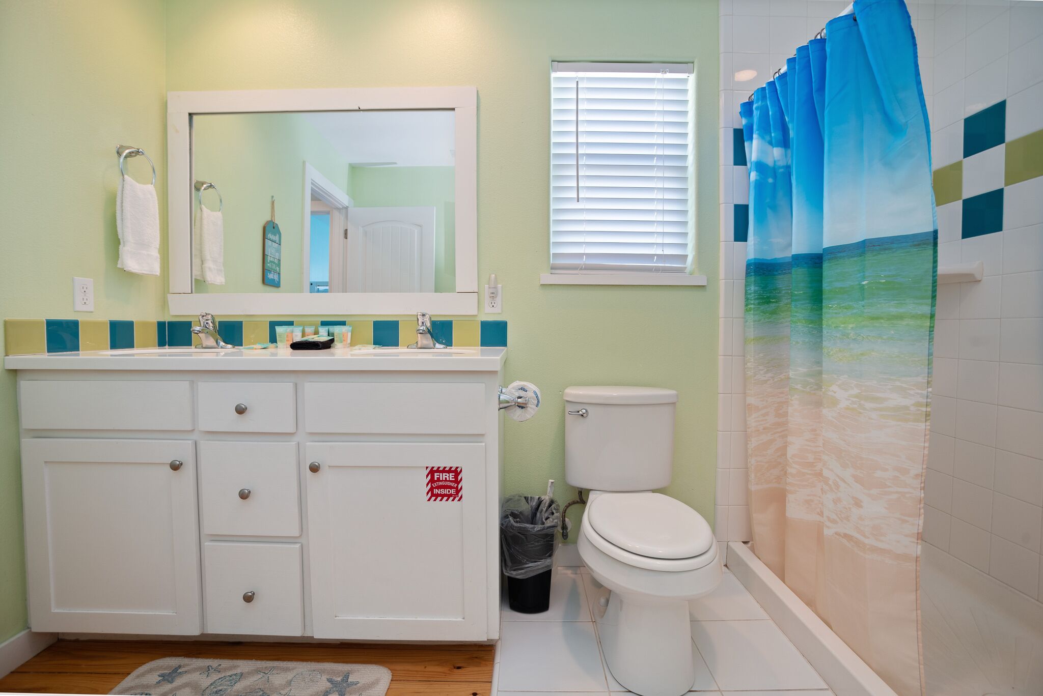 Une salle de bain avec un lavabo blanc, un miroir et des toilettes. Il y a une fenêtre avec des stores et un rideau de douche orné d’un motif de plage.