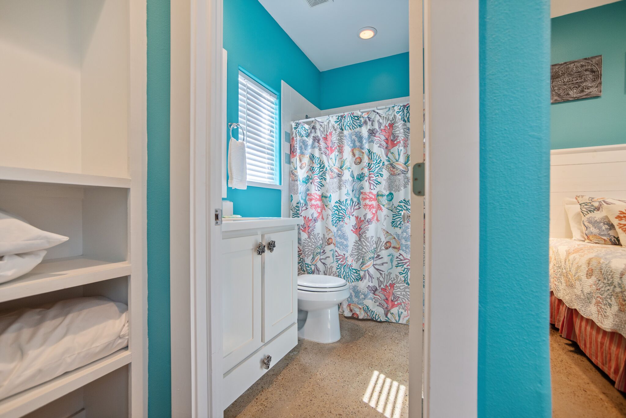 Une salle de bain avec des toilettes, un rideau de douche à motifs coralliens et, dans la pièce adjacente, un lit recouvert d’une couvre-lit à fleurs.