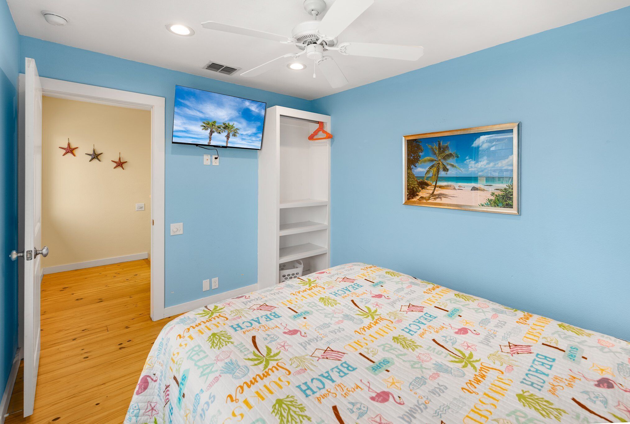 Un dormitorio con cama, ventilador de techo, televisor y un cuadro de una escena de playa.