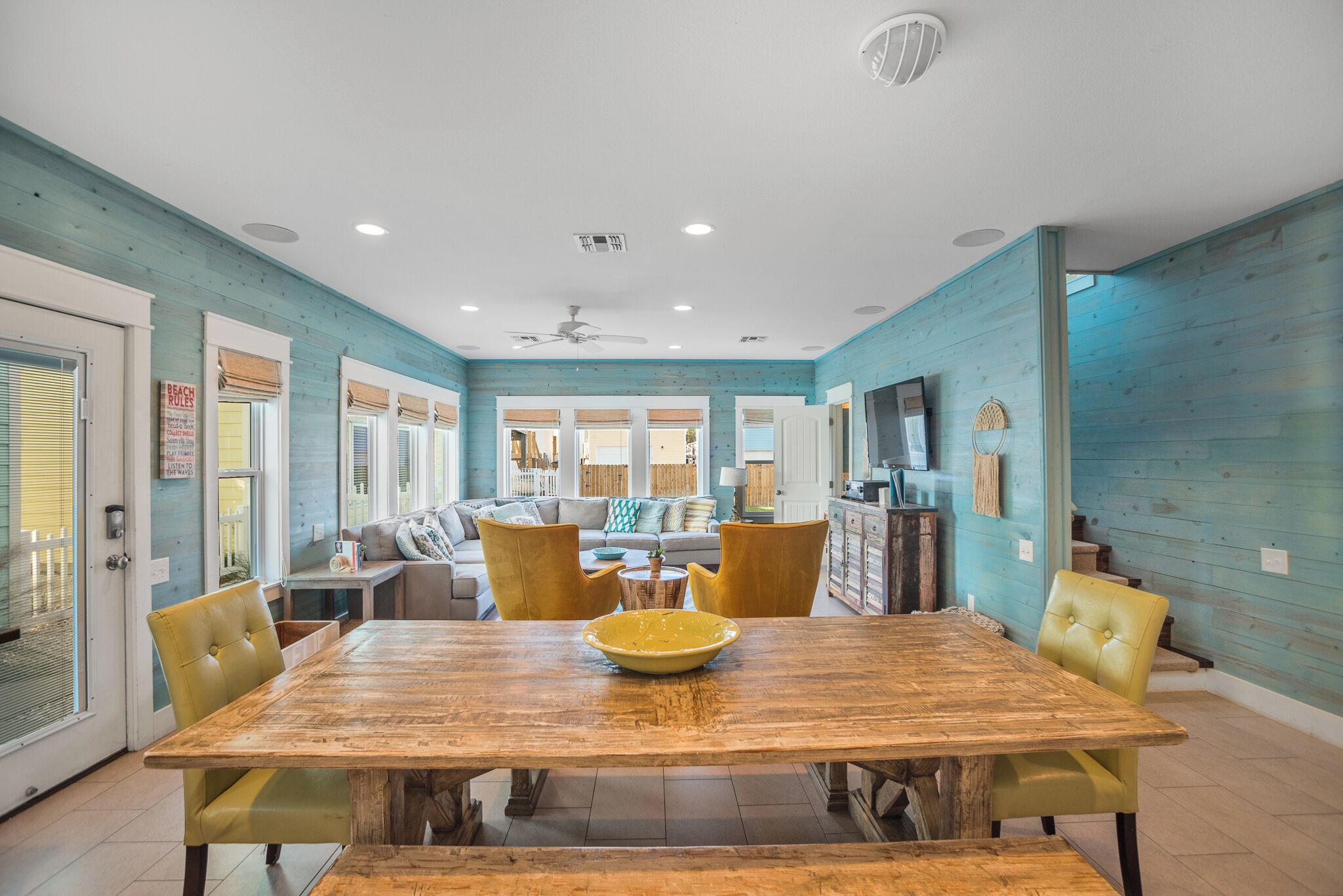 A spacious dining area with a wooden table and yellow chairs, a kitchen in the background, and a view of the outdoors through large windows.