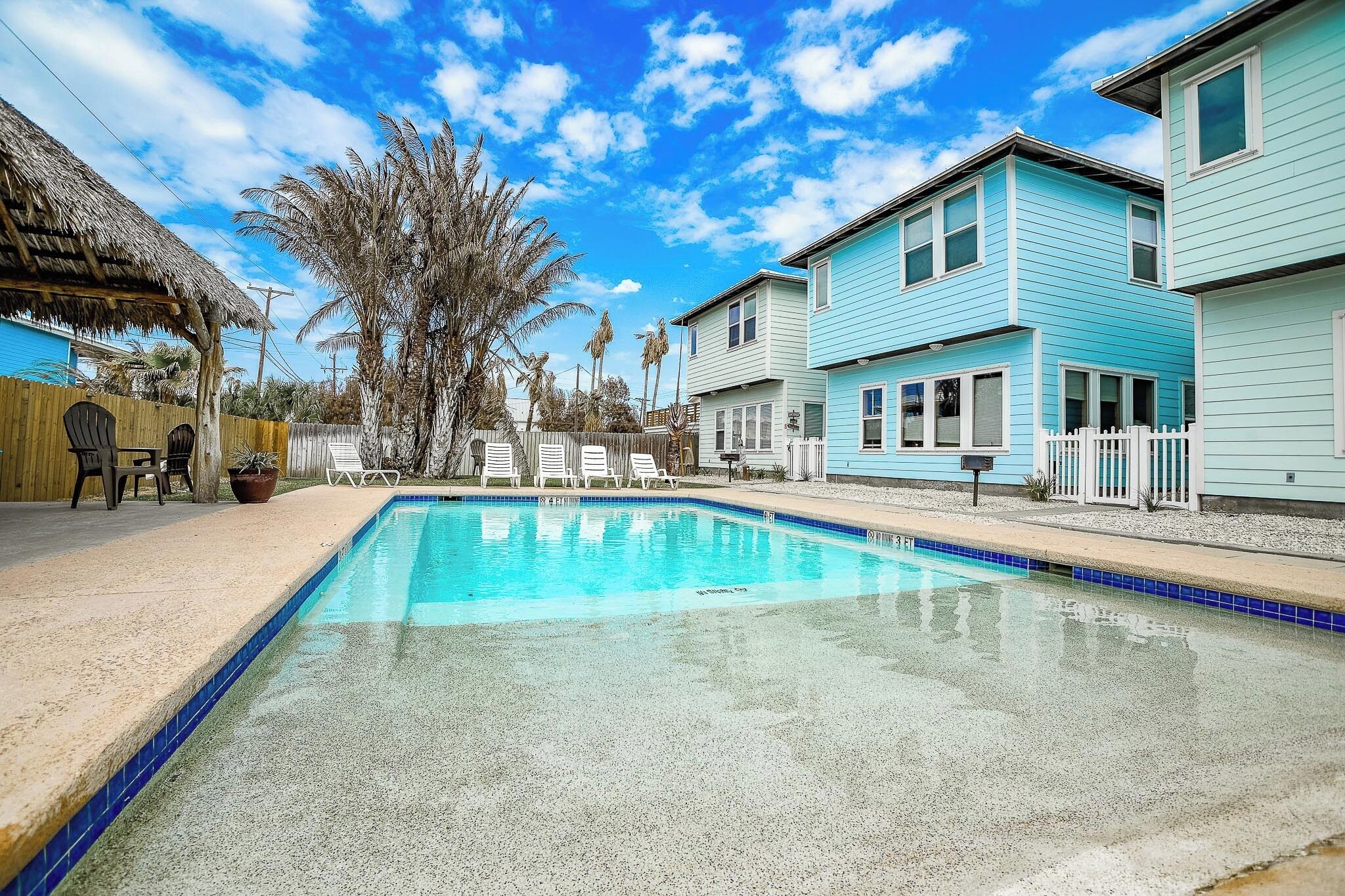 A swimming pool with lounge chairs, a wooden fence, and houses in the background.