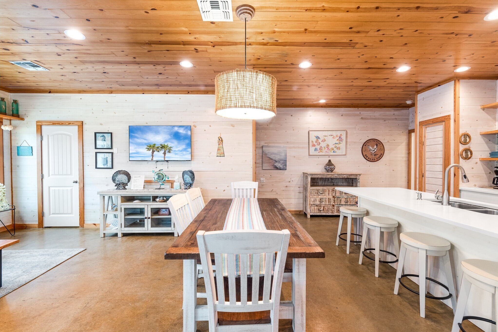 Teto de madeira, uma mesa de jantar com cadeiras, área de cozinha com pia e espaço para banquetes com banquinhos.