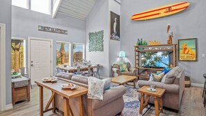 A spacious living room with a high ceiling, a dining area, and a kitchen. A surfboard is mounted on the wall, and there are various decorative items and a sign that reads "BEACH HOUSE".