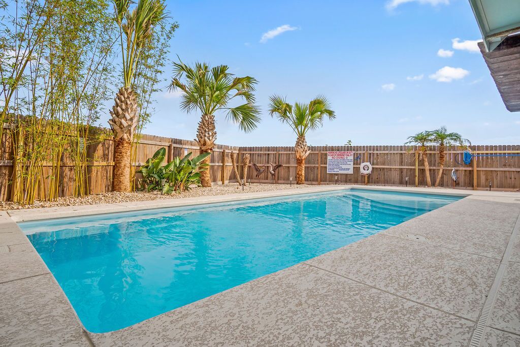 Ein rechteckiger Swimmingpool mit Holzzaun und Palmen im Hintergrund.