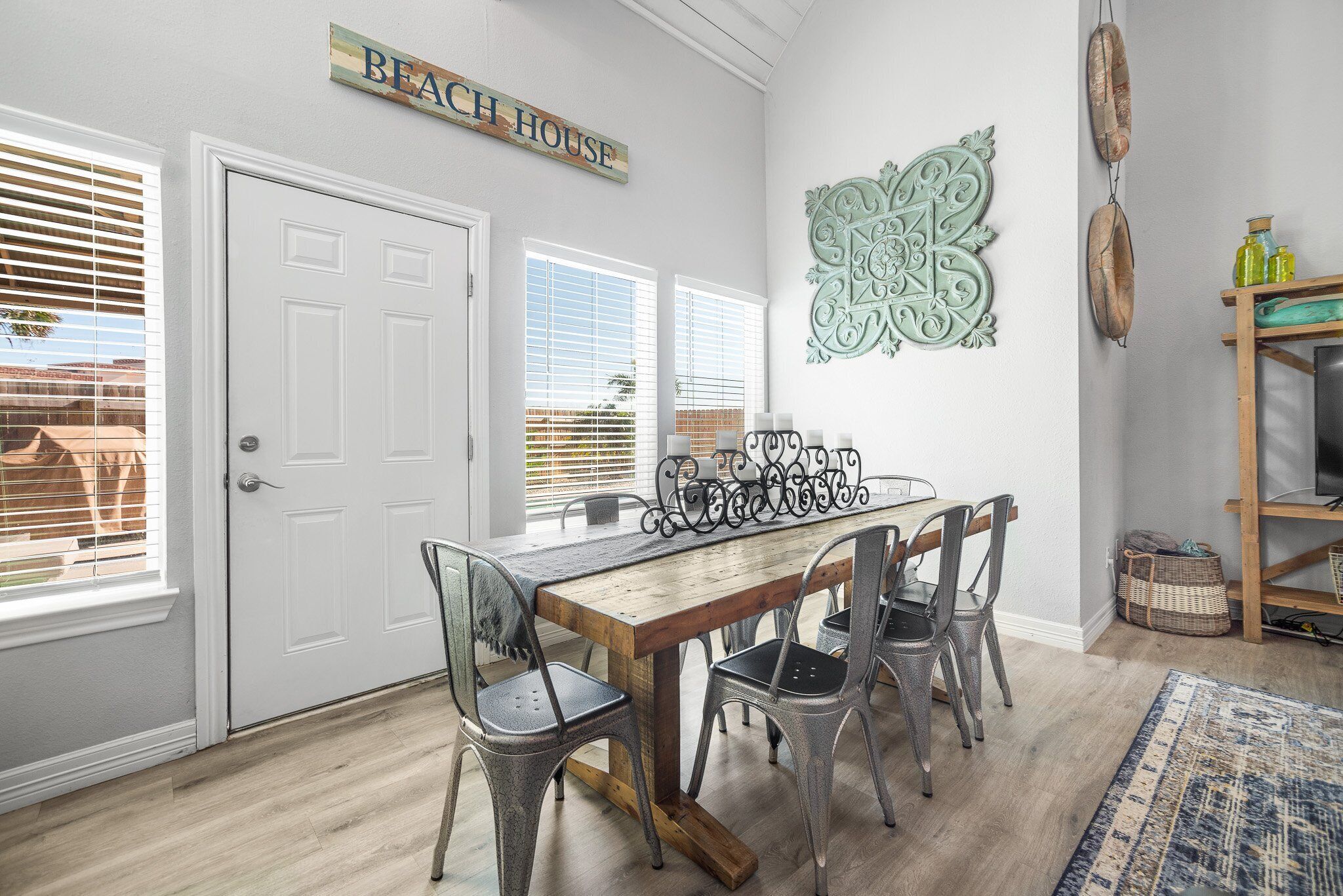 A dining area with a wooden table and metal chairs, a white door, and a sign that reads "BEACH HOUSE".