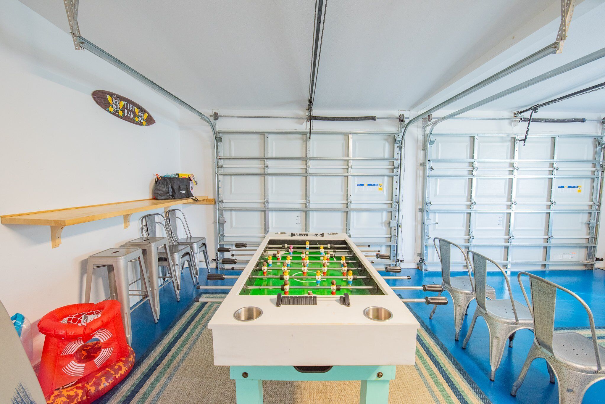 Een tafelvoetbaltafel in een ruimte met een blauwe vloer, metalen stoelen, een klein tafeltje met een tas en een bord op de muur.