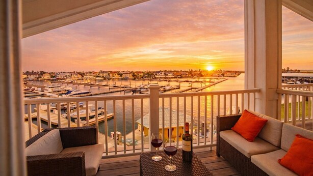 A balcony with wicker furniture, a table with wine glasses and a bottle, and a marina view during sunset. - Marina Del Bell - Elevator! Boat Slip for Rent! (Port Aransas)