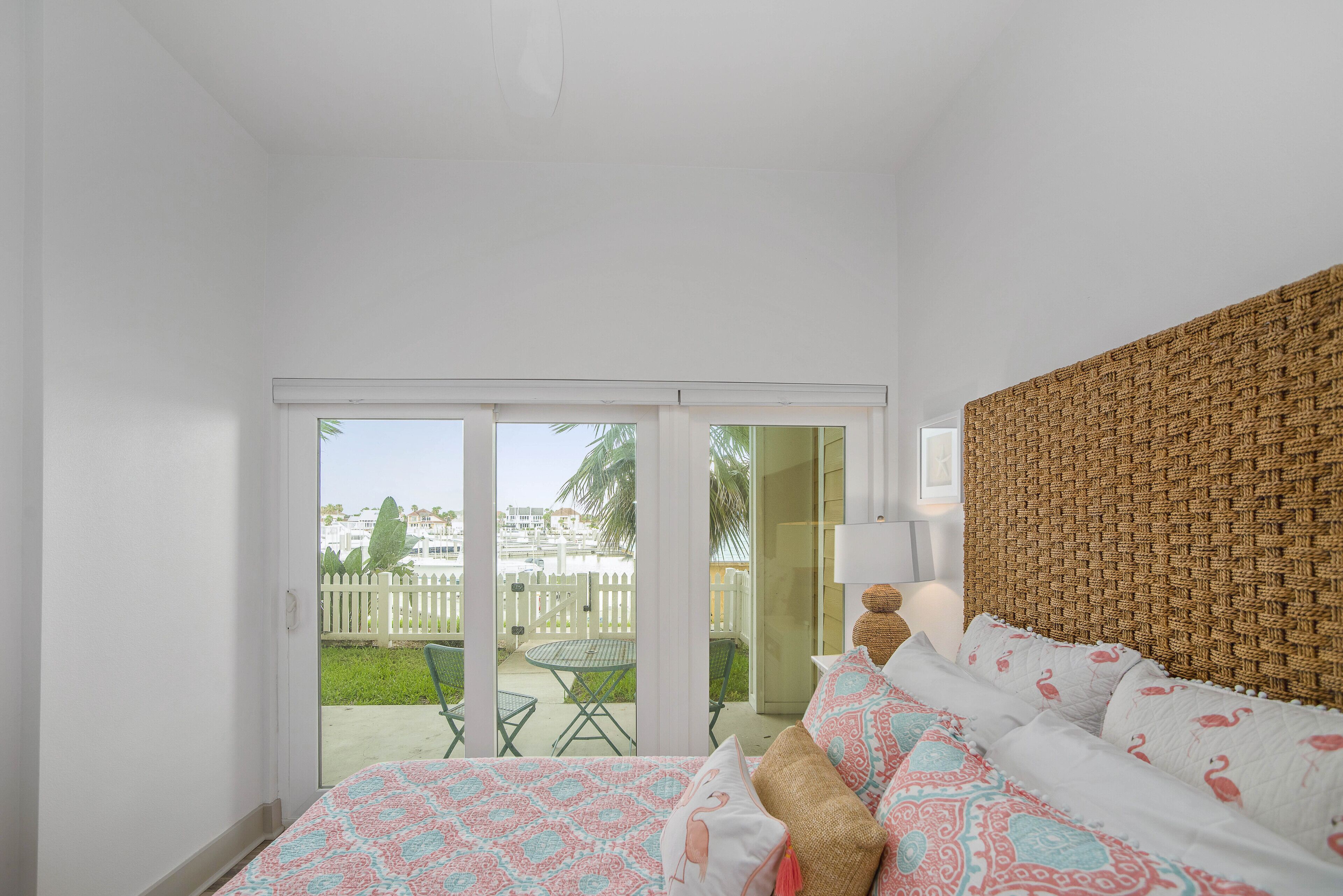 Ein Schlafzimmer mit Bett, einem gewobenen Kopfteil, Blick auf einen Balkon mit Tisch und Stühlen sowie einem weißen Zaun.