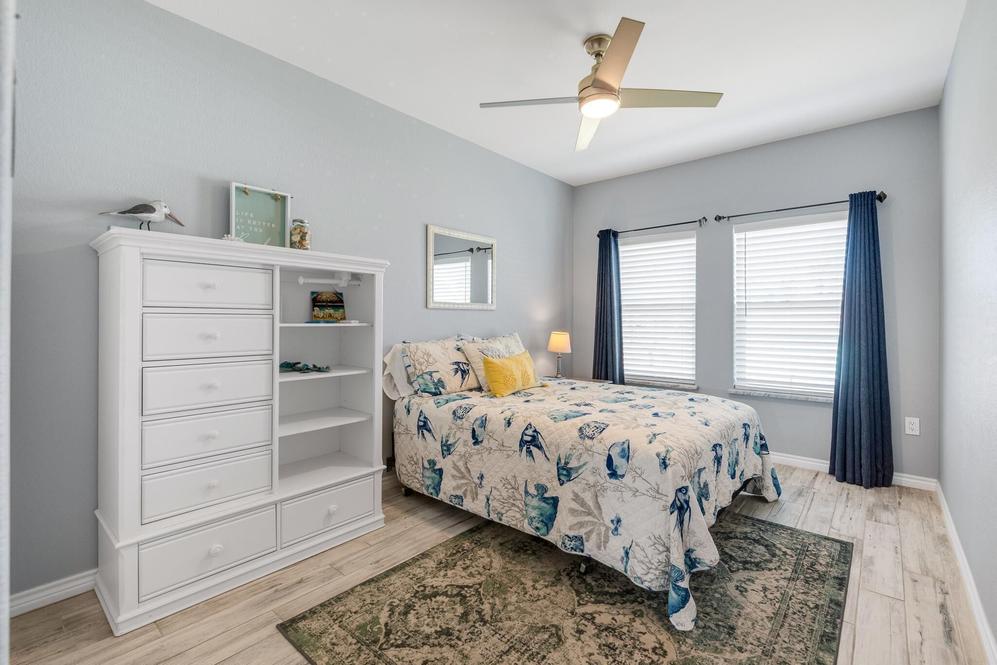 A bedroom with a bed, dresser, ceiling fan, windows with curtains, and a rug.