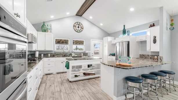 A modern kitchen with white cabinetry, a central island with stools, a clock on the wall, and a sign welcoming guests.