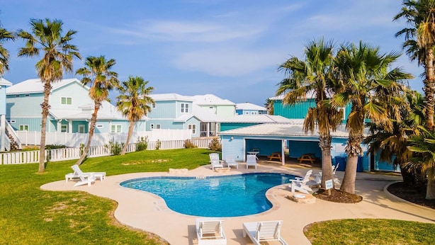 A swimming pool with lounge chairs, palm trees, and a building with a light blue exterior. - Beachside Bliss - Beach Gear Credit Included! (Port Aransas)
