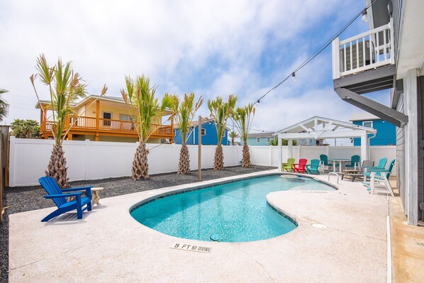 A swimming pool with a 5 ft no diving zone, surrounded by colorful Adirondack chairs, palm trees, and a building with a blue facade. - Latitude Adjustment - Private Pool/elevator! (Port Aransas)