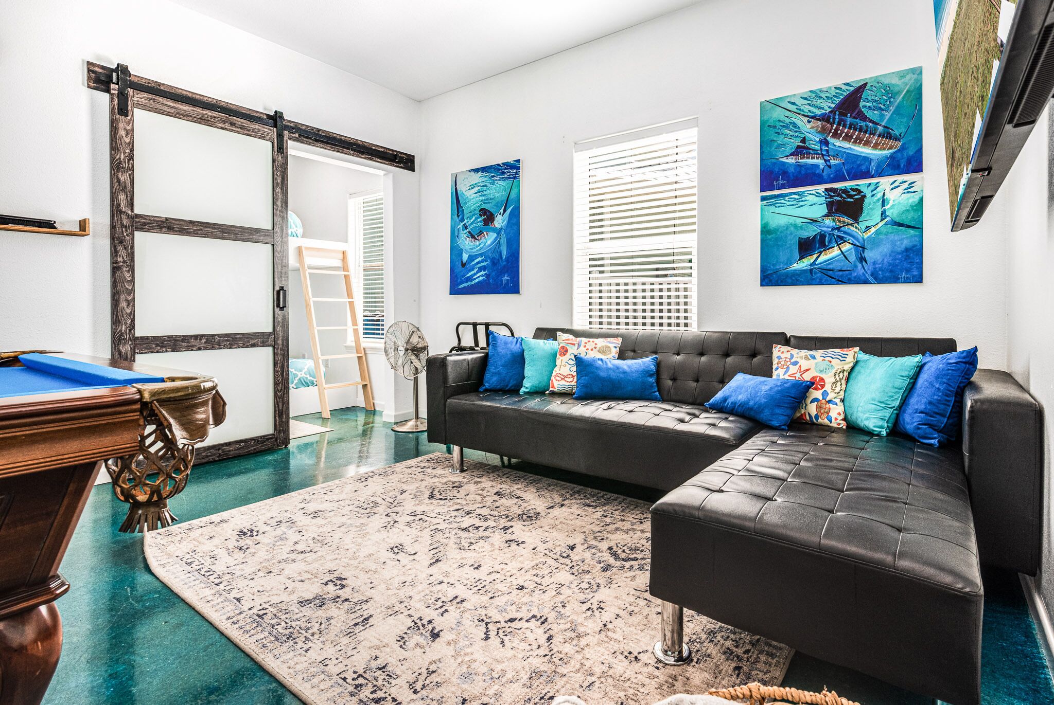 Ein Wohnzimmer mit Billardtisch, einer schwarzen Ledersofa mit bunten Kissen, einem großen abstrakten Gemälde und einer Schiebetür aus Glas.
