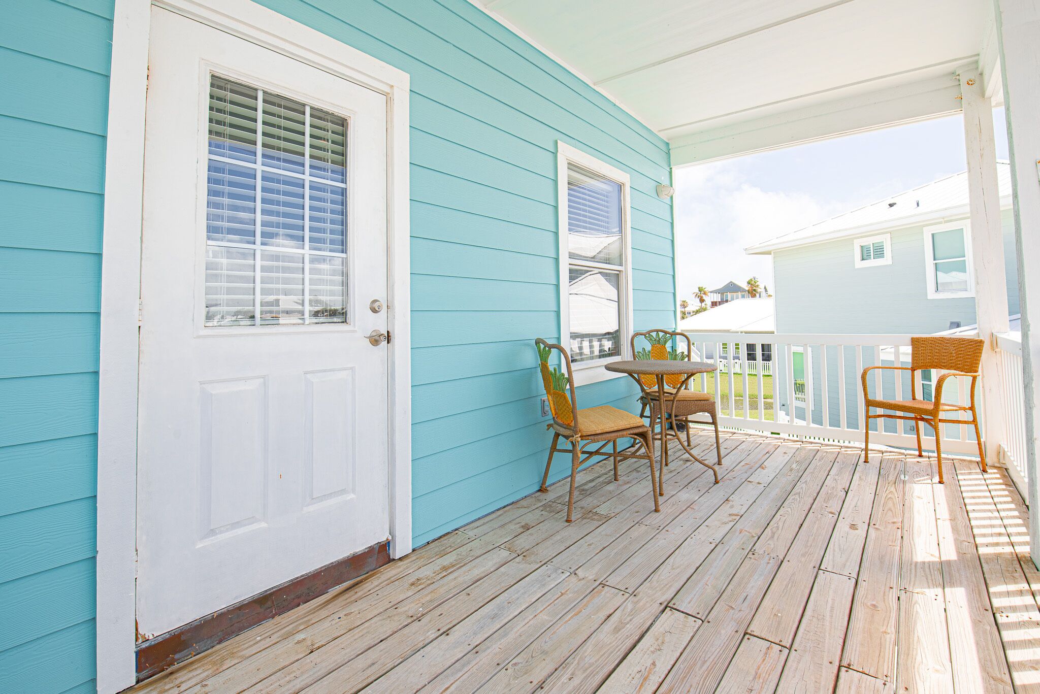 A wooden deck with a white door and a window with blinds, a table with chairs, and a view of another house in the background.
