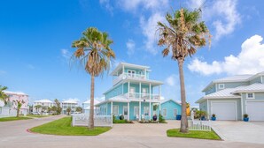 A two-story house with a balcony, surrounded by a white picket fence, palm trees, and a clear blue sky.