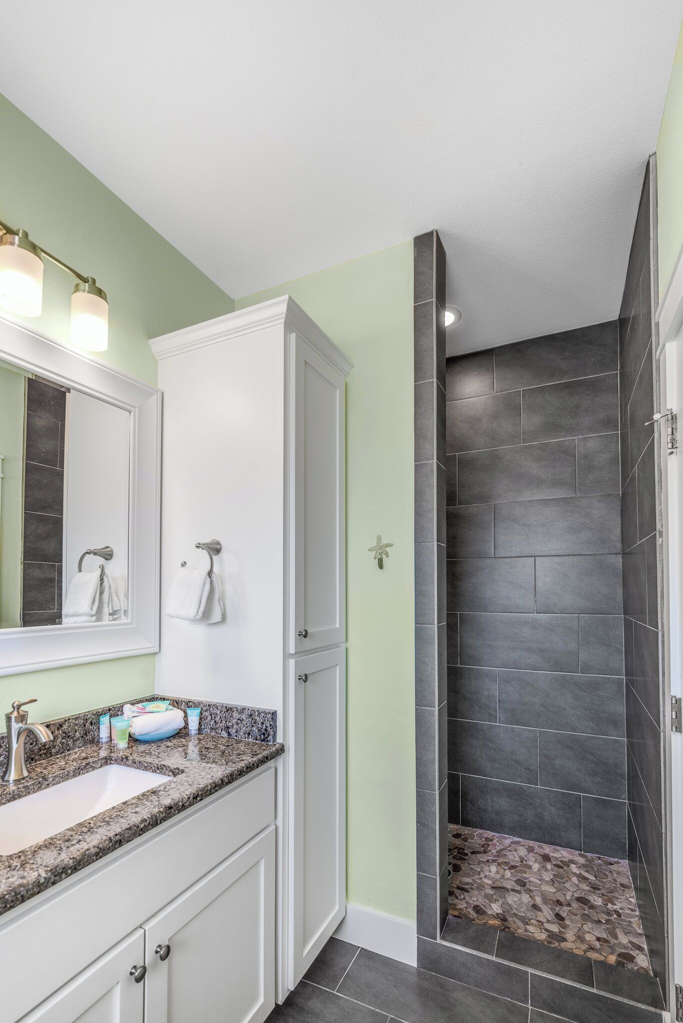 Une salle de bain avec un lavabo surmonté d’un plan de travail en granit, des meubles blancs, un grand miroir et une douche aux carreaux foncés.