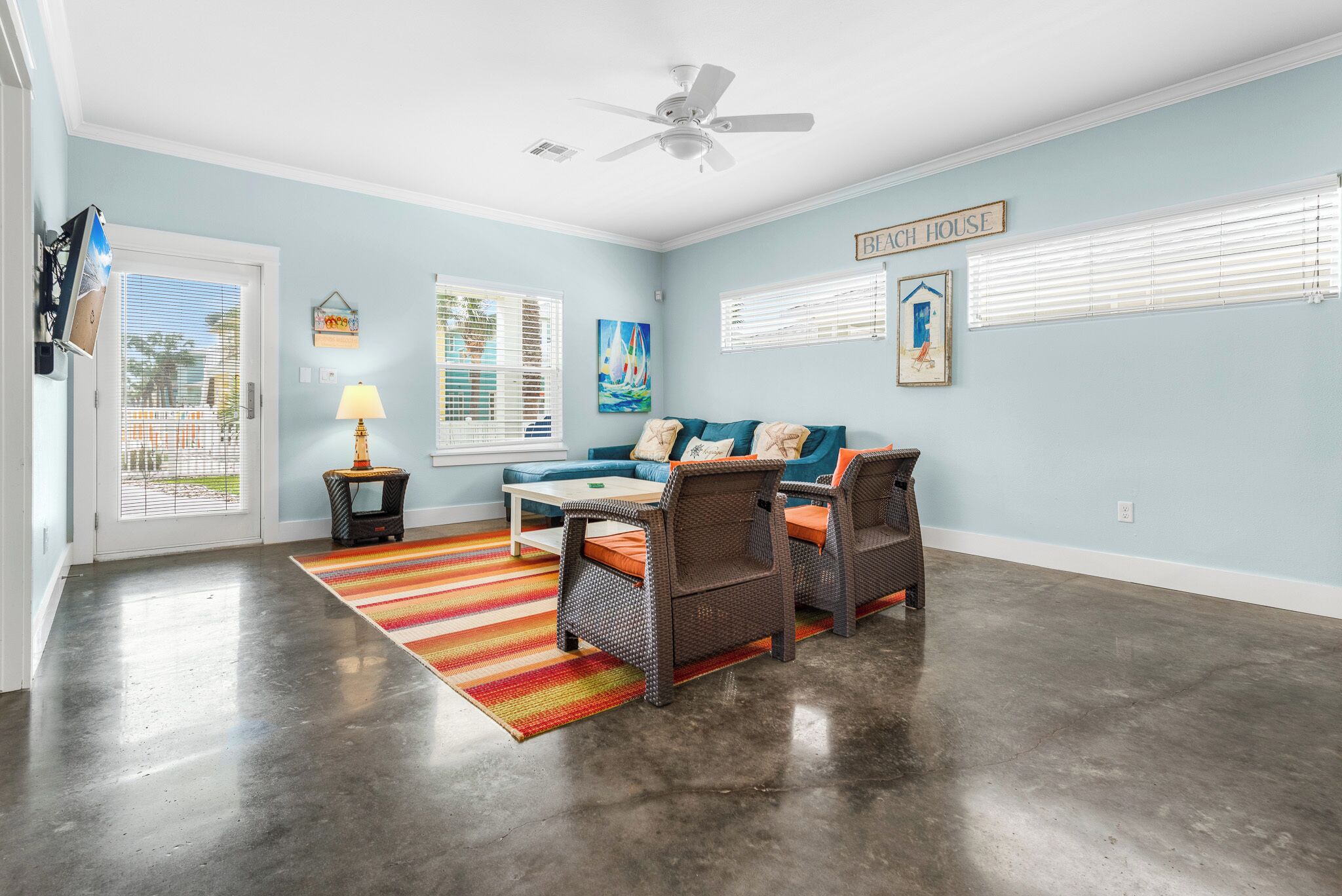 Ein geräumiges Wohnzimmer mit einem blauen Sofa, zwei Korbstühlen, einem Couchtisch, einem farbenfrohen Streifen Teppich, einem Deckenventilator und einem Schild mit der Aufschrift 'Beach House' an der Wand.