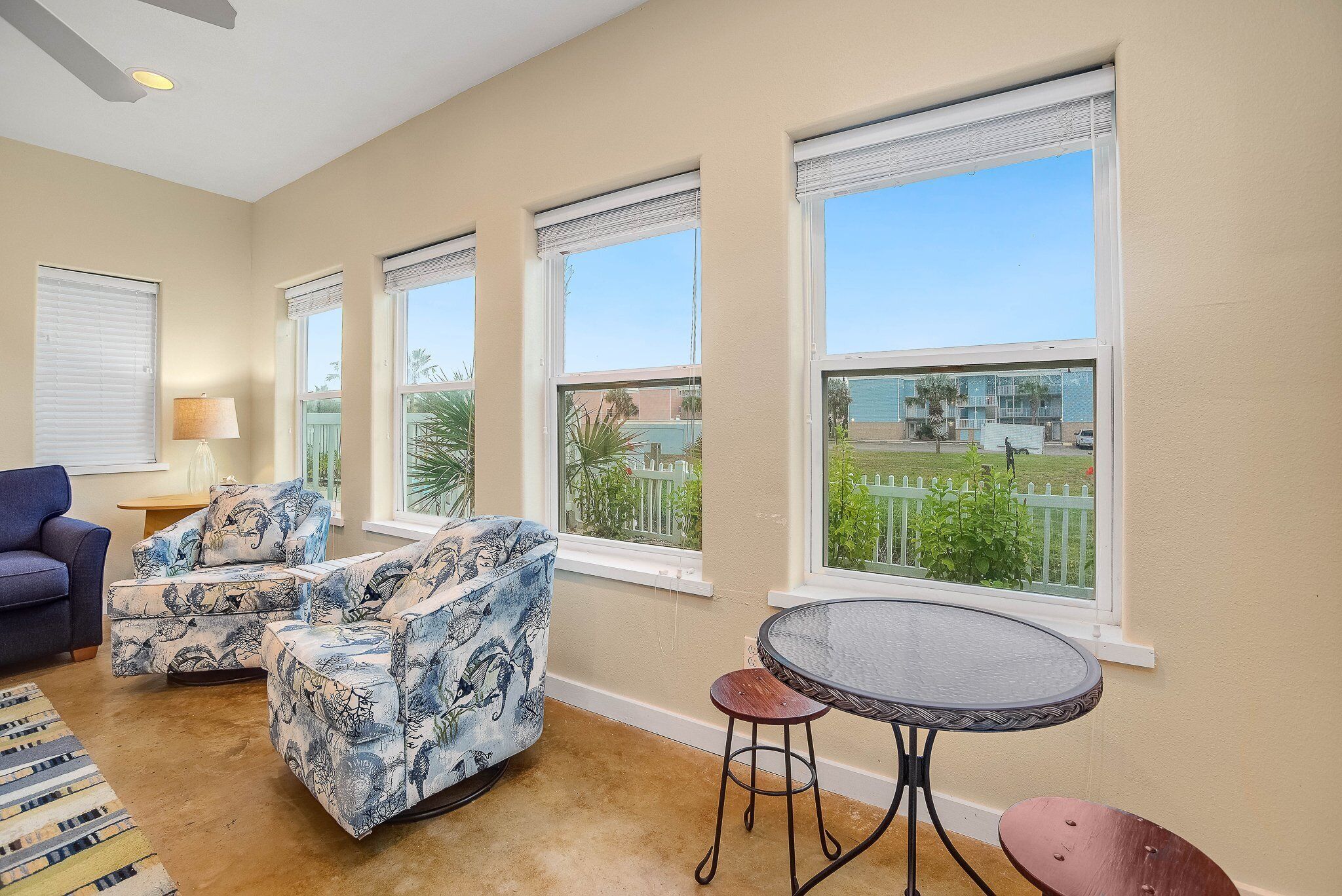 A living room with a sofa set, a small round table, and a chair. There are large windows with blinds, and a view of a fenced area outside.