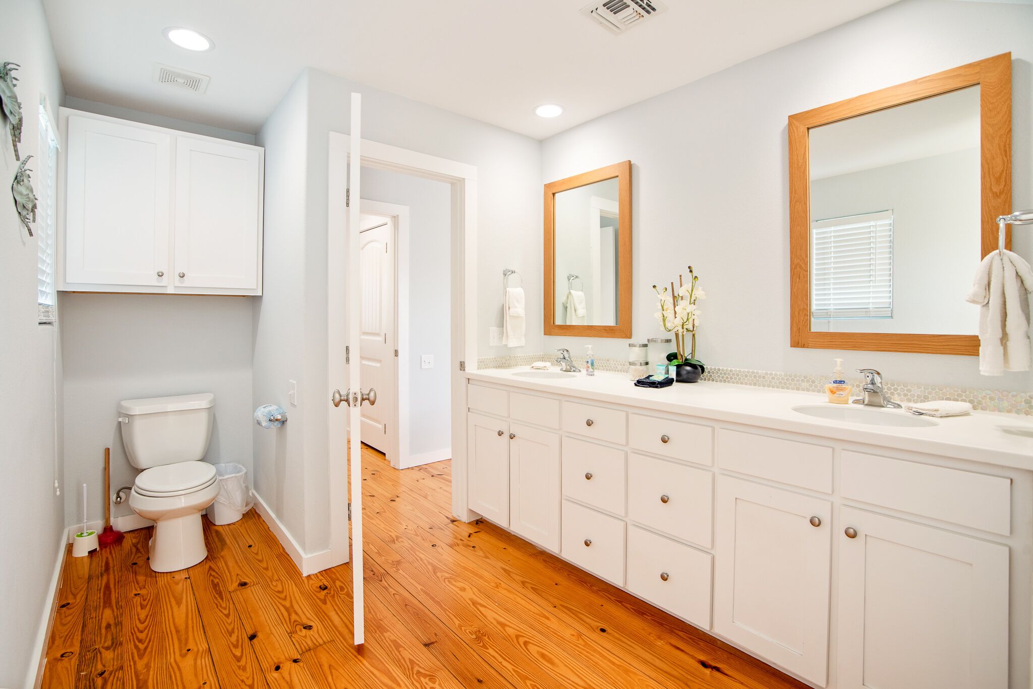 Een moderne badkamer met een houten vloer, witte badkamerkastjes, een grote spiegel en een toilet.