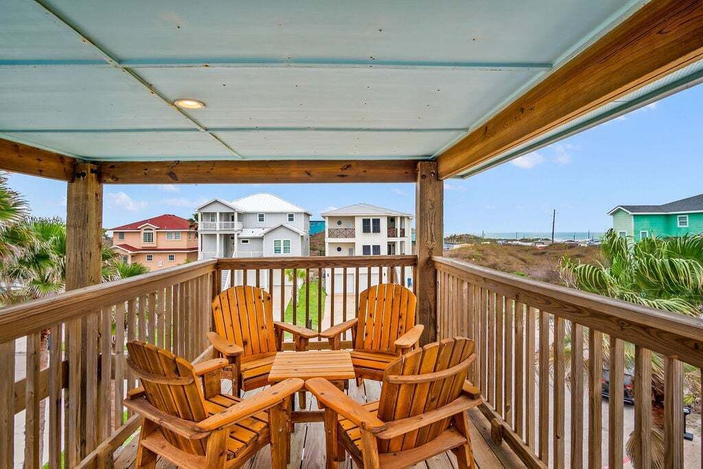 A wooden deck with four Adirondack chairs and a small table, overlooking a residential area with houses and a beach.
