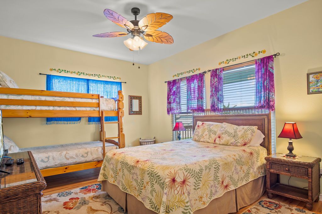 A bedroom with bunk beds, a ceiling fan, a bed with a floral bedspread, a nightstand with a lamp, and a window with purple curtains.