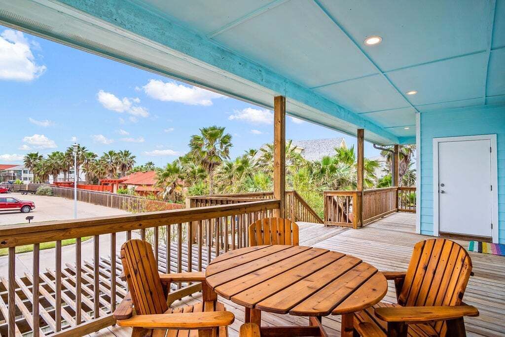 A wooden deck with a table and chairs, overlooking a street with palm trees and a red car.