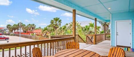 A wooden deck with a table and chairs, overlooking a street with palm trees and a red car.