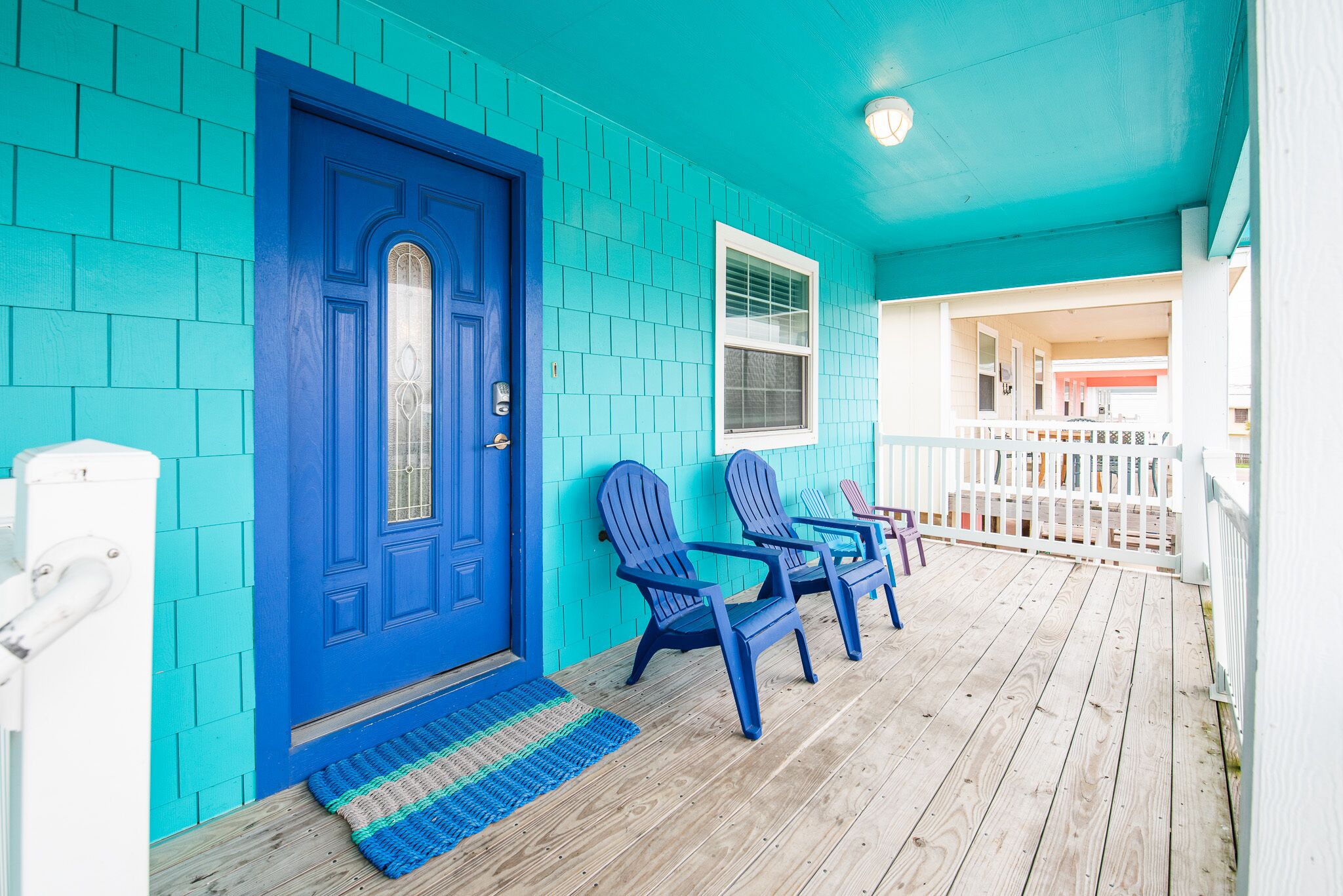 Fachada turquesa con una puerta azul, dos sillas Adirondack y una terraza de madera.