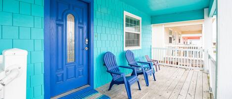 Fachada turquesa con una puerta azul, dos sillas Adirondack y una terraza de madera.