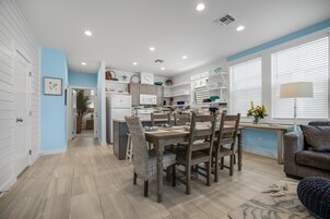 A modern kitchen with white cabinets, a dining area with a wooden table and chairs, and a living room with a sofa and a lamp.