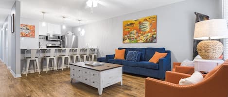 A modern living room with a blue sofa, orange armchair, and a coffee table. There is a kitchen area with white cabinets and a dining set.