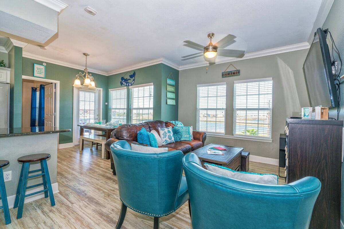 Un salon spacieux avec un canapé en cuir, deux fauteuils bleus, une table basse en bois et un ventilateur de plafond.