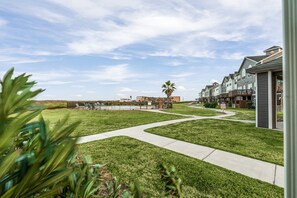 A residential area with a pathway leading to a body of water, surrounded by houses with balconies and a palm tree.